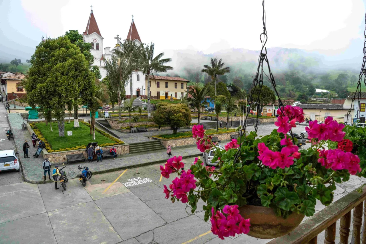 Plaza central de San José de la Montaña con la iglesia de dos torres, palmas, casas de colores y montañas cubiertas de niebla. En primer plano, una matera con geranios fucsia en un balcón.
